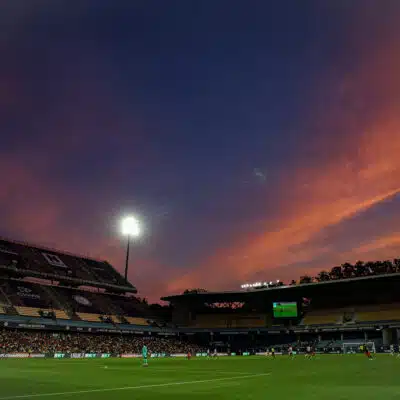 Soccer stadium at dusk with orange clouds, bright floodlights, and players on a green pitch.