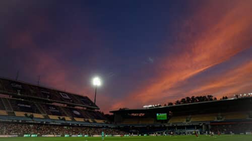 Soccer stadium at dusk with orange clouds, bright floodlights, and players on a green pitch.