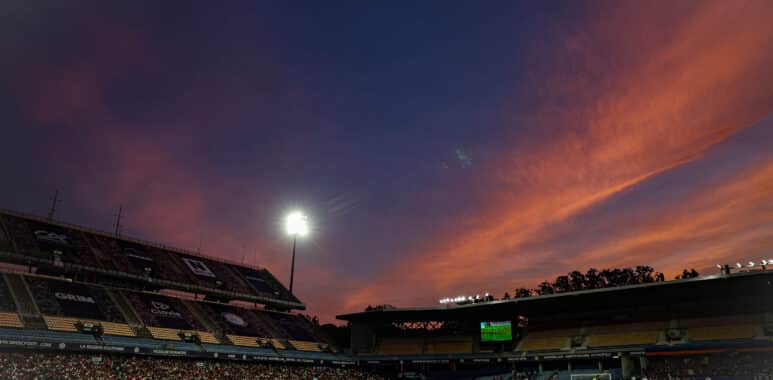 Soccer stadium at dusk with orange clouds, bright floodlights, and players on a green pitch.