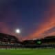 Soccer stadium at dusk with orange clouds, bright floodlights, and players on a green pitch.