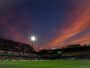 Soccer stadium at dusk with orange clouds, bright floodlights, and players on a green pitch.