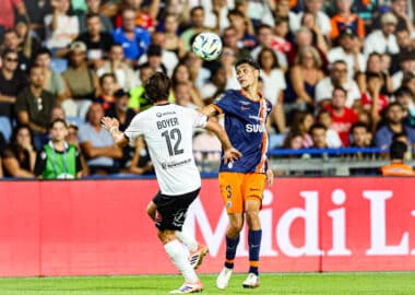 Two soccer players in mid-air contesting a header as a ball hovers above them, stadium crowd in the background.