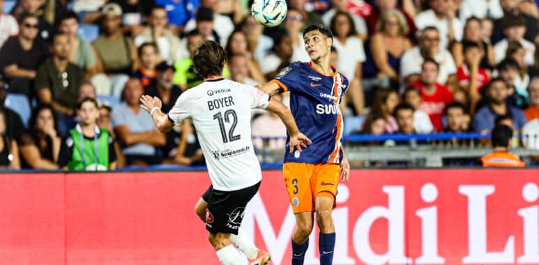 Two soccer players in mid-air contesting a header as a ball hovers above them, stadium crowd in the background.