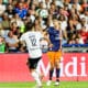 Two soccer players in mid-air contesting a header as a ball hovers above them, stadium crowd in the background.