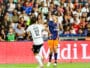 Two soccer players in mid-air contesting a header as a ball hovers above them, stadium crowd in the background.