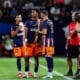 Soccer players in blue-orange uniforms standing on the field with coaches and officials nearby during a match.