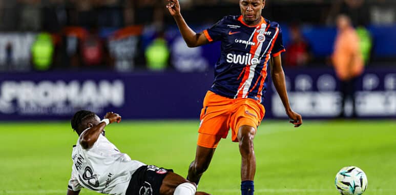 Soccer match moment: a blue-and-orange team player stands over a sliding white-jersey defender as the ball rolls nearby.
