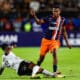 Soccer match moment: a blue-and-orange team player stands over a sliding white-jersey defender as the ball rolls nearby.