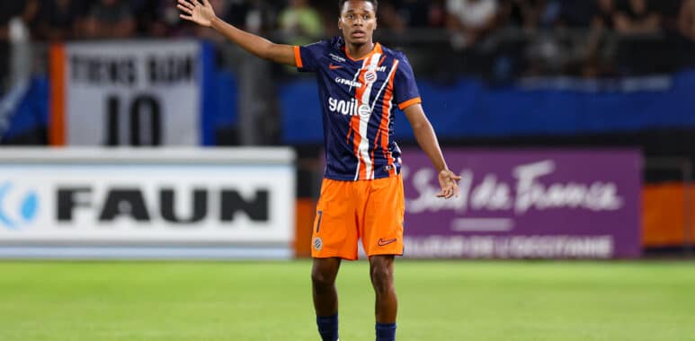Soccer player in blue-orange kit raises left arm on a green field with blurred stadium crowd behind him, signaling or calling for the ball.