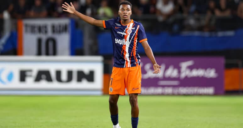 Soccer player in blue-orange kit raises left arm on a green field with blurred stadium crowd behind him, signaling or calling for the ball.