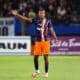 Soccer player in blue-orange kit raises left arm on a green field with blurred stadium crowd behind him, signaling or calling for the ball.