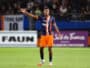 Soccer player in blue-orange kit raises left arm on a green field with blurred stadium crowd behind him, signaling or calling for the ball.