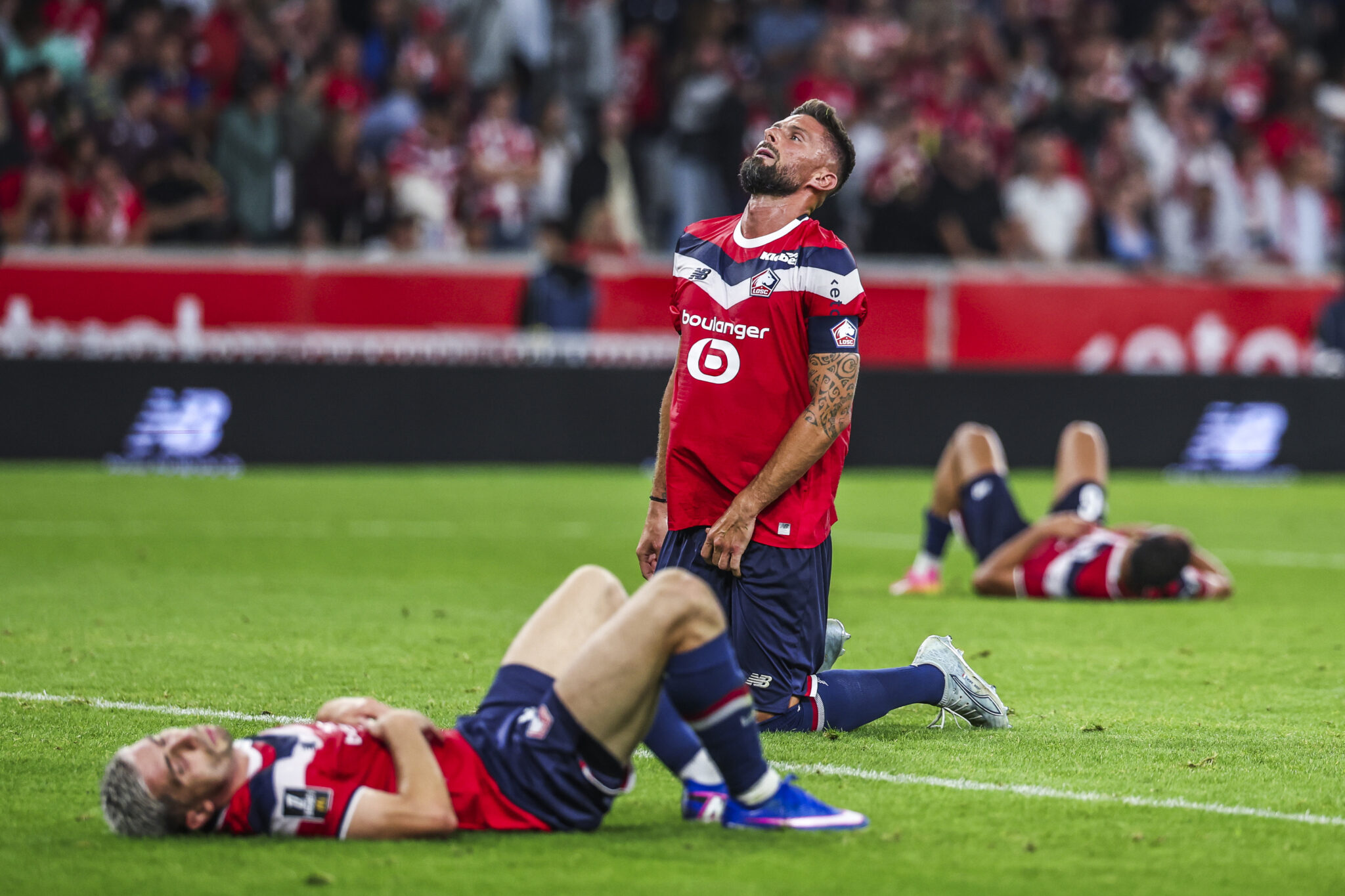 Giroud Lille OSC v Paris Saint-Germain - Ligue 1 McDonalds 20262027 | AllezPaillade.com Soccer players in red jerseys on a green field, with one standing and others lying on the turf after a collision, crowd of spectators in the stands behind them.