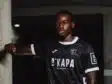 Portrait of a Black male soccer player in a black jersey with white trim, leaning against a concrete column in a dim indoor setting.