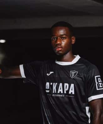 Portrait of a Black male soccer player in a black jersey with white trim, leaning against a concrete column in a dim indoor setting.