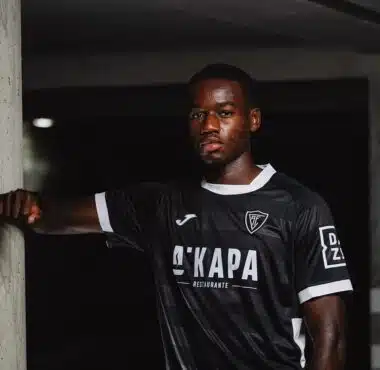 Portrait of a Black male soccer player in a black jersey with white trim, leaning against a concrete column in a dim indoor setting.