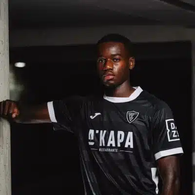Portrait of a Black male soccer player in a black jersey with white trim, leaning against a concrete column in a dim indoor setting.