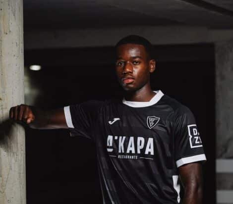 Portrait of a Black male soccer player in a black jersey with white trim, leaning against a concrete column in a dim indoor setting.