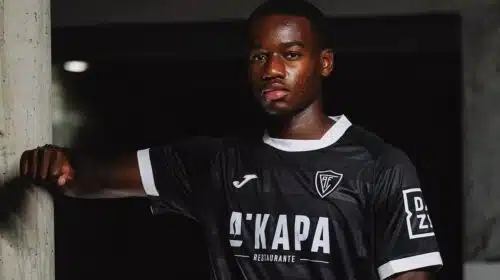 Portrait of a Black male soccer player in a black jersey with white trim, leaning against a concrete column in a dim indoor setting.