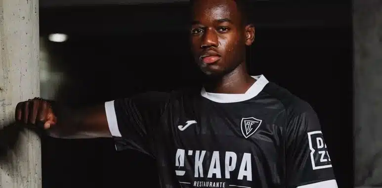 Screenshot | AllezPaillade.com Portrait of a Black male soccer player in a black jersey with white trim, leaning against a concrete column in a dim indoor setting.