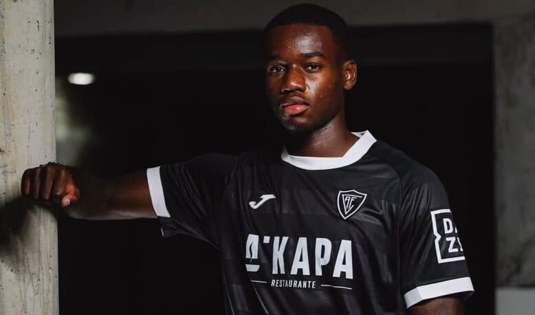 Portrait of a Black male soccer player in a black jersey with white trim, leaning against a concrete column in a dim indoor setting.
