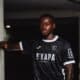 Portrait of a Black male soccer player in a black jersey with white trim, leaning against a concrete column in a dim indoor setting.