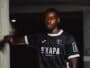 Portrait of a Black male soccer player in a black jersey with white trim, leaning against a concrete column in a dim indoor setting.
