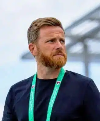 Man with a reddish beard wearing a black shirt and a green lanyard, standing outdoors and looking to the right.