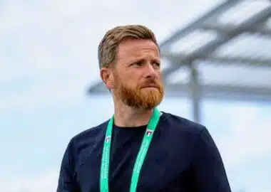 Man with a reddish beard wearing a black shirt and a green lanyard, standing outdoors and looking to the right.