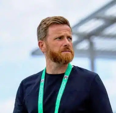 Man with a reddish beard wearing a black shirt and a green lanyard, standing outdoors and looking to the right.