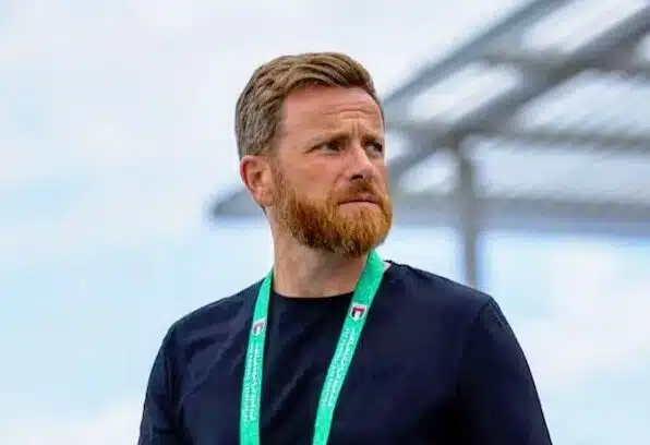 Man with a reddish beard wearing a black shirt and a green lanyard, standing outdoors and looking to the right.
