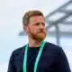 Man with a reddish beard wearing a black shirt and a green lanyard, standing outdoors and looking to the right.