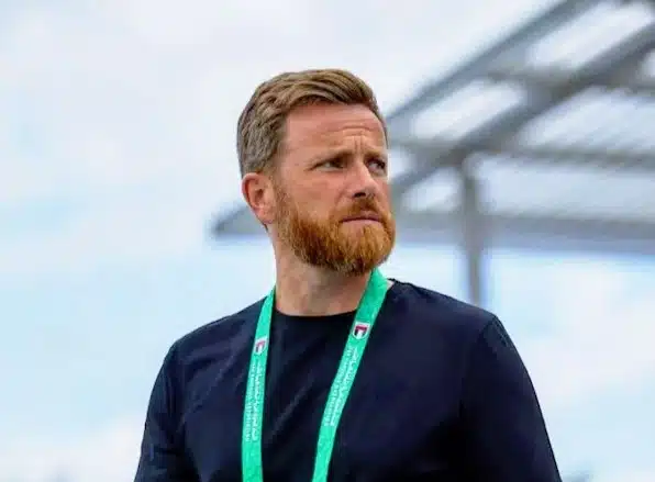 Man with a reddish beard wearing a black shirt and a green lanyard, standing outdoors and looking to the right.