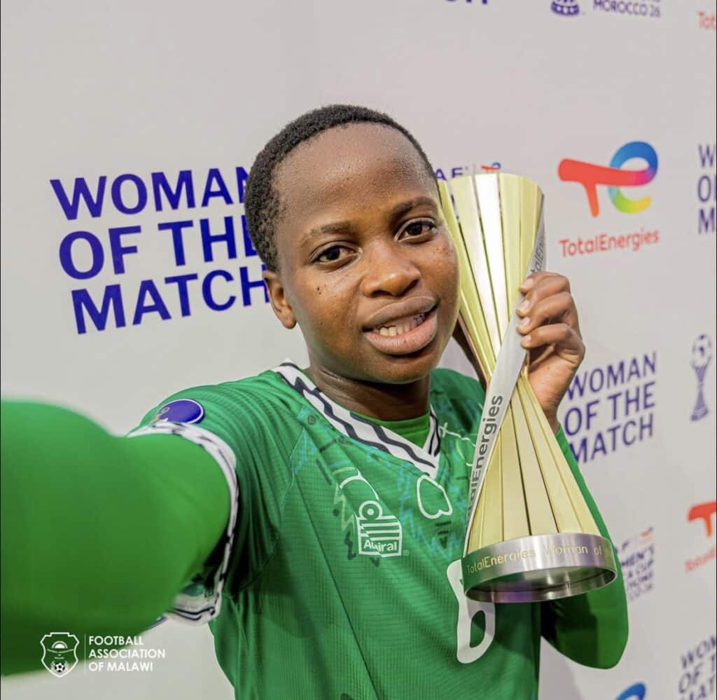 Young female soccer player in a green jersey holds a tall gold trophy, posing for a selfie with a backdrop reading'Woman of the Match' and sponsor logos.