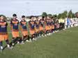 Youth soccer players stand in a long line on a green pitch before kickoff, wearing orange-and-navy uniforms while opponents in white-and-black stripes wait behind them. A few referees in bright yellow jerseys stand nearby.