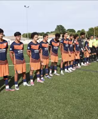 Screenshot | AllezPaillade.com Youth soccer players stand in a long line on a green pitch before kickoff, wearing orange-and-navy uniforms while opponents in white-and-black stripes wait behind them. A few referees in bright yellow jerseys stand nearby.