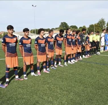 Youth soccer players stand in a long line on a green pitch before kickoff, wearing orange-and-navy uniforms while opponents in white-and-black stripes wait behind them. A few referees in bright yellow jerseys stand nearby.