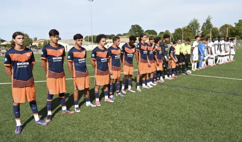 Youth soccer players stand in a long line on a green pitch before kickoff, wearing orange-and-navy uniforms while opponents in white-and-black stripes wait behind them. A few referees in bright yellow jerseys stand nearby.