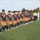 Youth soccer players stand in a long line on a green pitch before kickoff, wearing orange-and-navy uniforms while opponents in white-and-black stripes wait behind them. A few referees in bright yellow jerseys stand nearby.