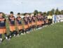 Youth soccer players stand in a long line on a green pitch before kickoff, wearing orange-and-navy uniforms while opponents in white-and-black stripes wait behind them. A few referees in bright yellow jerseys stand nearby.