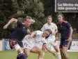 Female soccer players contest the ball during a match on a grassy field, white vs navy/orange kits in a tight challenge.