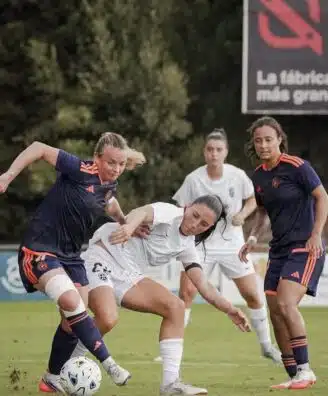 Female soccer players contest the ball during a match on a grassy field, white vs navy/orange kits in a tight challenge.
