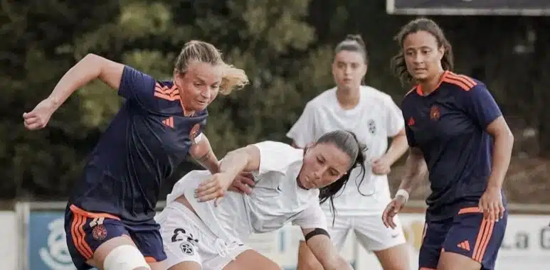 Female soccer players contest the ball during a match on a grassy field, white vs navy/orange kits in a tight challenge.