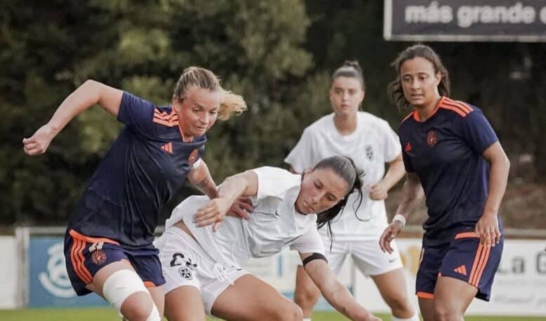Female soccer players contest the ball during a match on a grassy field, white vs navy/orange kits in a tight challenge.
