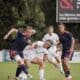 Female soccer players contest the ball during a match on a grassy field, white vs navy/orange kits in a tight challenge.