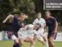 Female soccer players contest the ball during a match on a grassy field, white vs navy/orange kits in a tight challenge.