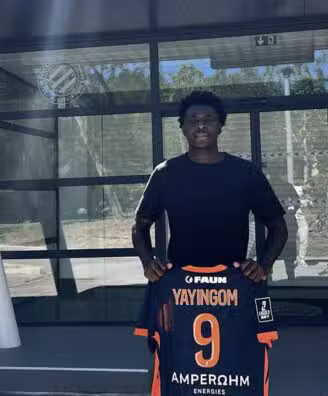 Young man standing outside a glass building, holding a dark sports jersey with orange trim and the name YAYINGOM number 9 on the back/front edge visible.