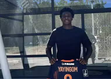 Young man standing outside a glass building, holding a dark sports jersey with orange trim and the name YAYINGOM number 9 on the back/front edge visible.