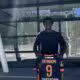 Young man standing outside a glass building, holding a dark sports jersey with orange trim and the name YAYINGOM number 9 on the back/front edge visible.