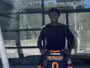 Young man standing outside a glass building, holding a dark sports jersey with orange trim and the name YAYINGOM number 9 on the back/front edge visible.